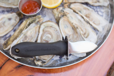 Oyster Shucker knife with a black handle that says 'Charleston Shucker Co.' in black letters, on a plate of oysters.