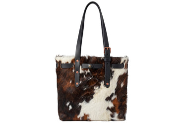 A structured cowhide tote bag with black leather shoulder straps and trim, featuring a brown and white natural cow print pattern and copper-toned hardware, shown against a plain white background.