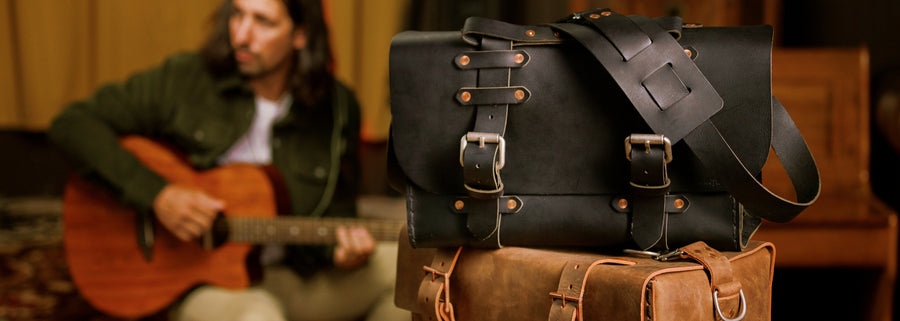 No. 4311 Satchel in Black stacked on top of a duffle bag with a man playing the guitar in the background. 